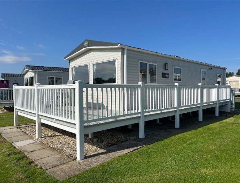 A white static caravan with a large white deck sits on a grassy lawn under a clear blue sky at Golden Sands Holiday Park. A perfect base for Perfect base for dog-friendly activities in Lincolnshire.