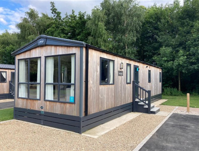 A stylish static caravan with grey steps and large windows, set against a backdrop of trees at Kingfisher Caravan Park. Perfect base for dog-friendly activities in Lincolnshire