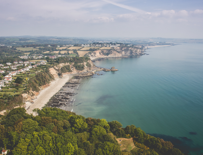 A beautiful view of steep cliffs leading to a sandy beach by the calm sea, with trees and houses on top of the cliffs.