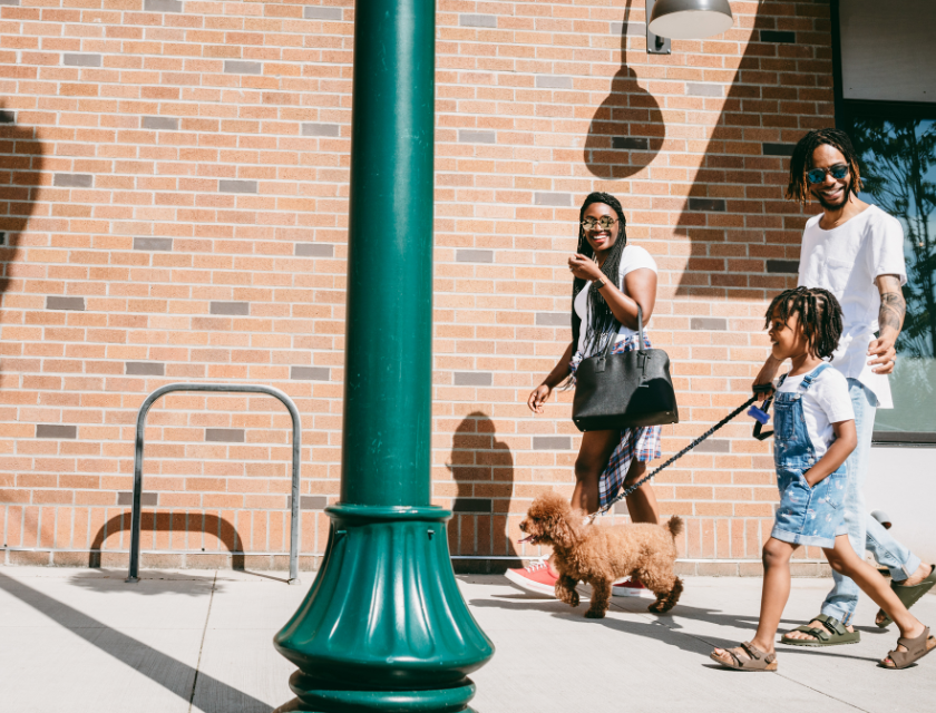 Family including a young child holding the leash of a small, curly-haired dog, walking along a sunny sidewalk next to a brick wall.