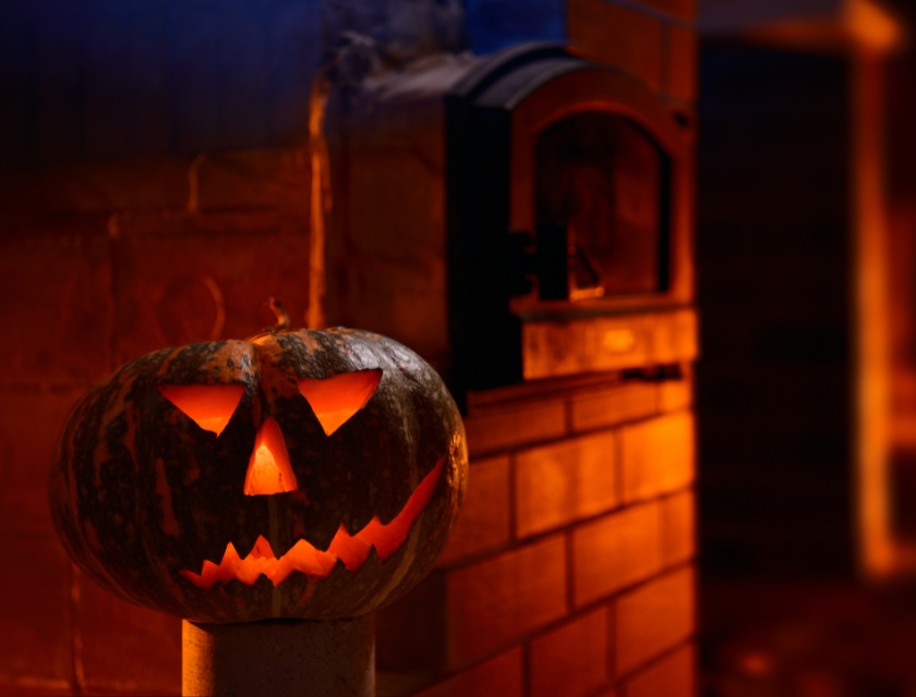 A carved pumpkin glowing with a spooky face, placed in front of a warm brick oven at night.