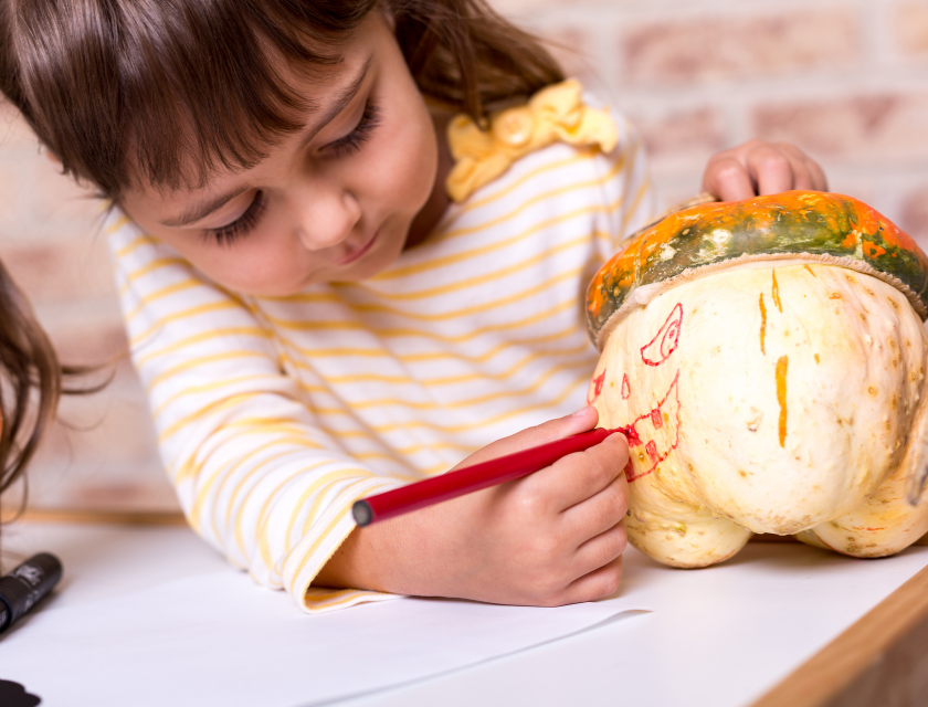 A young girl drawing a face on a pumpkin with a red marker, preparing for Halloween.