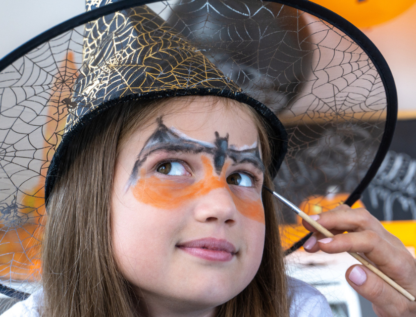 A girl wearing a witch hat having her face painted with a bat design for Halloween.