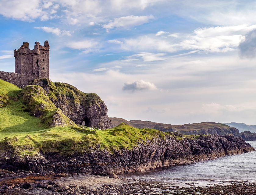 A castle ruin on top of cliffs, with the sea below and hills in the background.