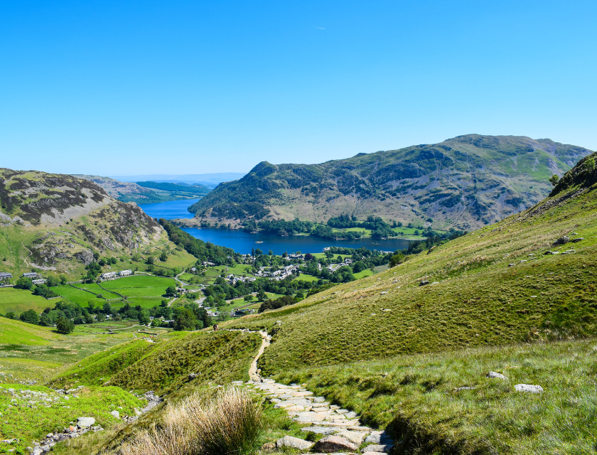 Path leading down a green valley to Ullswater Lake in the Lake District, with rolling hills. Treat yourself to a Lake District trip to see some of the best views while on an autumn walk in the UK. 