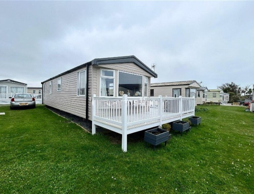 A beige mobile static caravan with a white deck and a car parked beside it, set in a grassy area on a cloudy day. Taken at Harlyn Sands Holiday Park, a perfect base for dog-friendly activities in Cornwall.