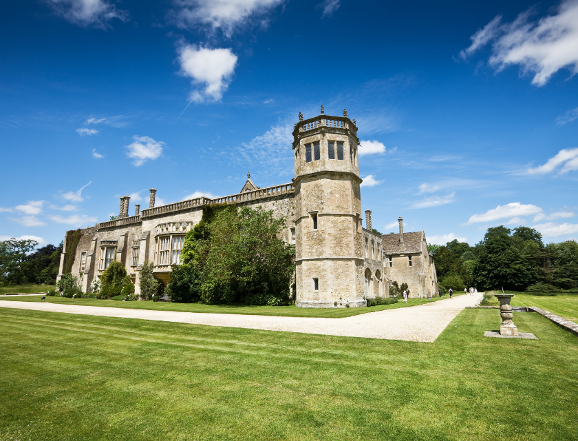 Lacock Abbey in Wiltshire on a sunny day. The historic building and lush green lawn are visible.