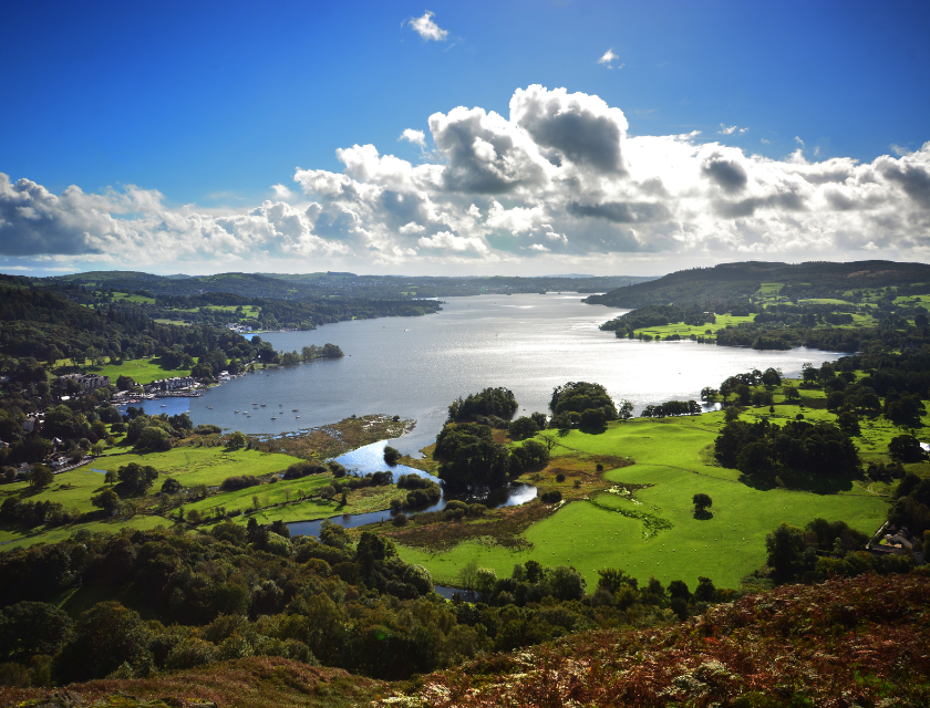 A sunny view over Lake Windermere in the Lake District, showing green fields and hills meeting the clear, calm water.