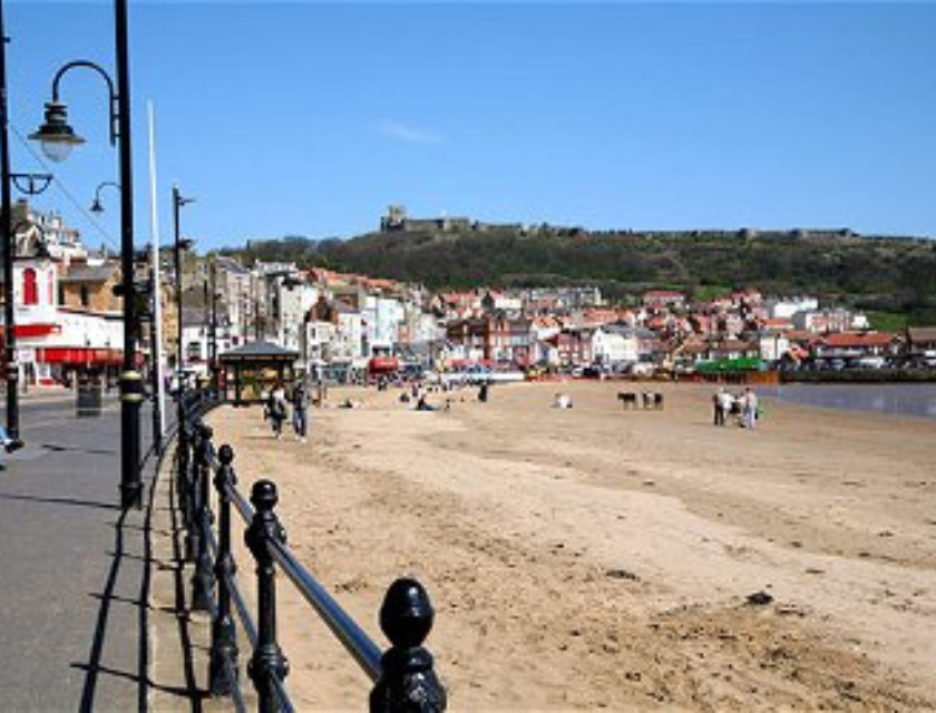 A coastal view of a bustling seaside town with a sandy beach, promenade, and colourful shops, offering a perfect spot for a holiday break.