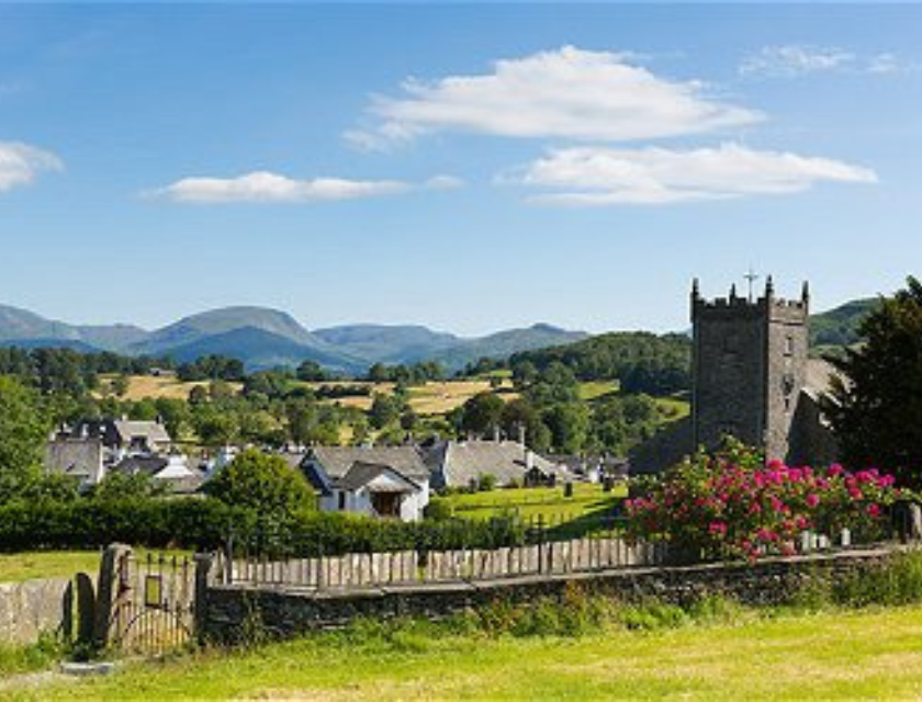 English countryside village with a stone church, surrounded by lush greenery and hills, with mountains under a bright blue sky.