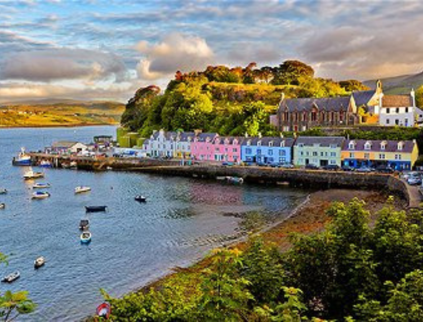 A picturesque Scottish harbour town with rows of brightly coloured houses along the water, surrounded by lush greenery and reflecting calm waters.