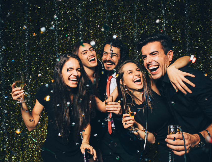 A group of five friends in party attire, laughing and holding champagne glasses, surrounded by confetti and a sparkling backdrop for New Year's Eve.