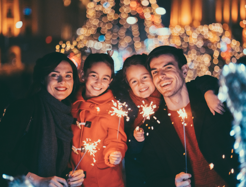 A family of four smiling and holding sparklers in front of a glowing, festive background, enjoying the magic of New Year's Eve together.