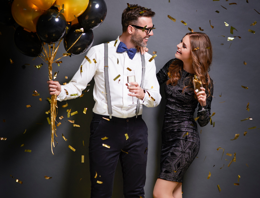 A well-dressed couple celebrating New Year's Eve, holding black and gold balloons, with confetti falling around them.