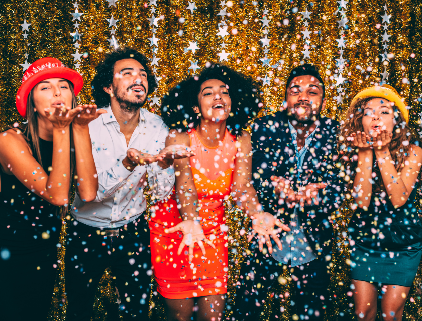 A group of friends dressed for New Year's Eve, celebrating with confetti and posing in front of a shimmering gold backdrop.