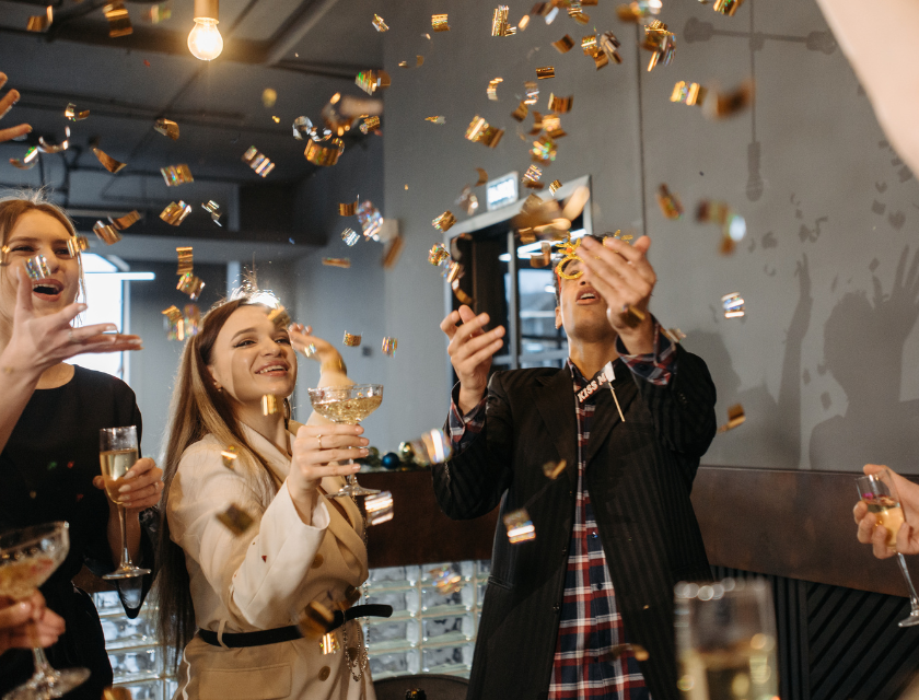 A lively New Year's Eve party indoors, with guests throwing confetti and raising glasses of champagne in a festive setting.