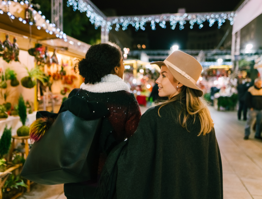 Two women smiling as they walk through a Christmas market filled with festive decorations and lights.