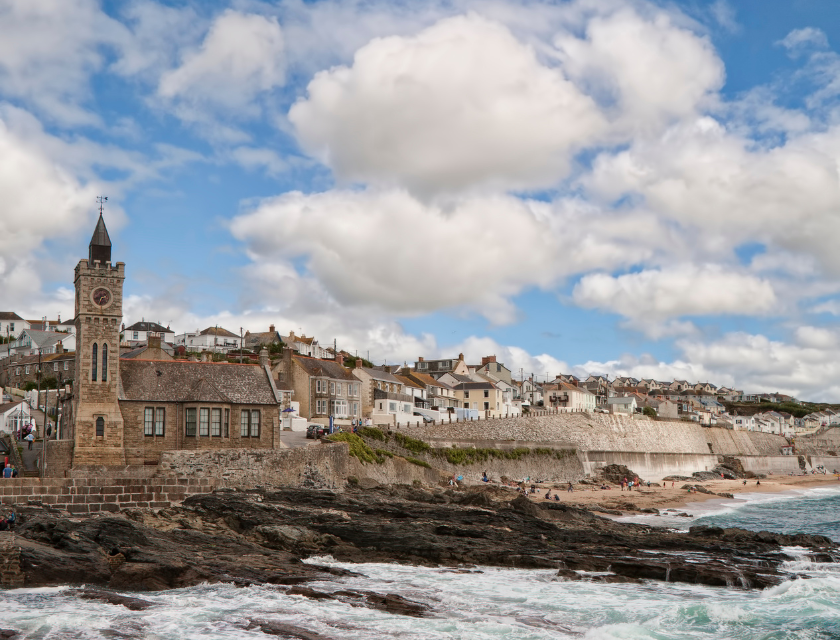 Coastal town of Porthleven, Cornwall, showing its stone clock tower and rocky coastline & a cloudy sky.