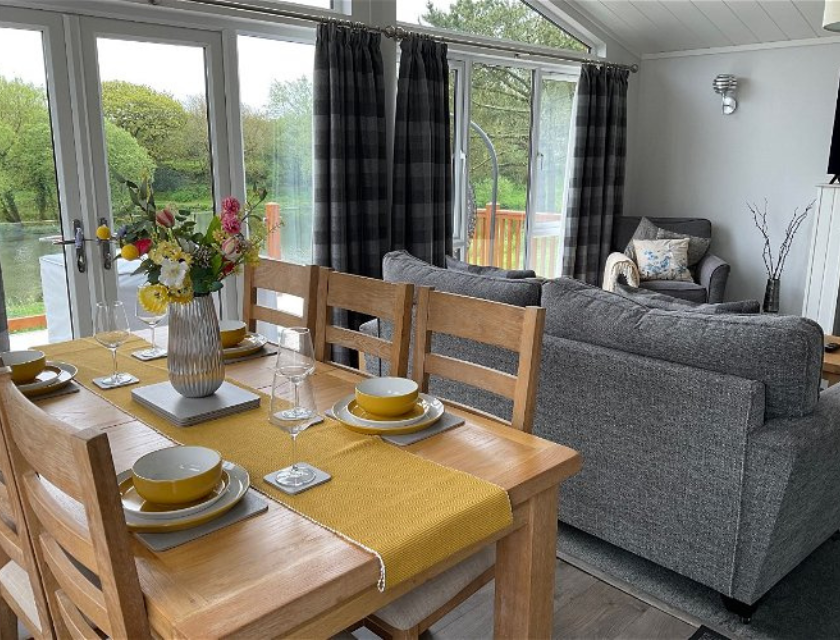 Dining room with a table set for four, a sofa, and large windows looking out to a lake and trees. Stay at White Acres Holiday Park during your visits to Cornwalls Christmas markets. 