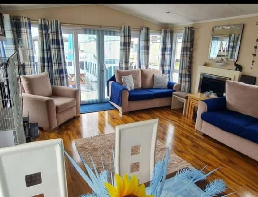 Static caravan living room at Lyons Robin Hood in North Wales with sofas with blue cushions & throws. There are large windows bringing in light from outside.