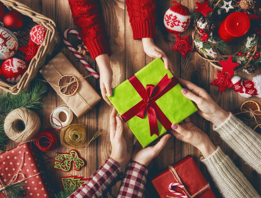 Family exchanging a gift wrapped in green paper with a red bow, surrounded by Christmas decorations, candles, and festive wrapping supplies.