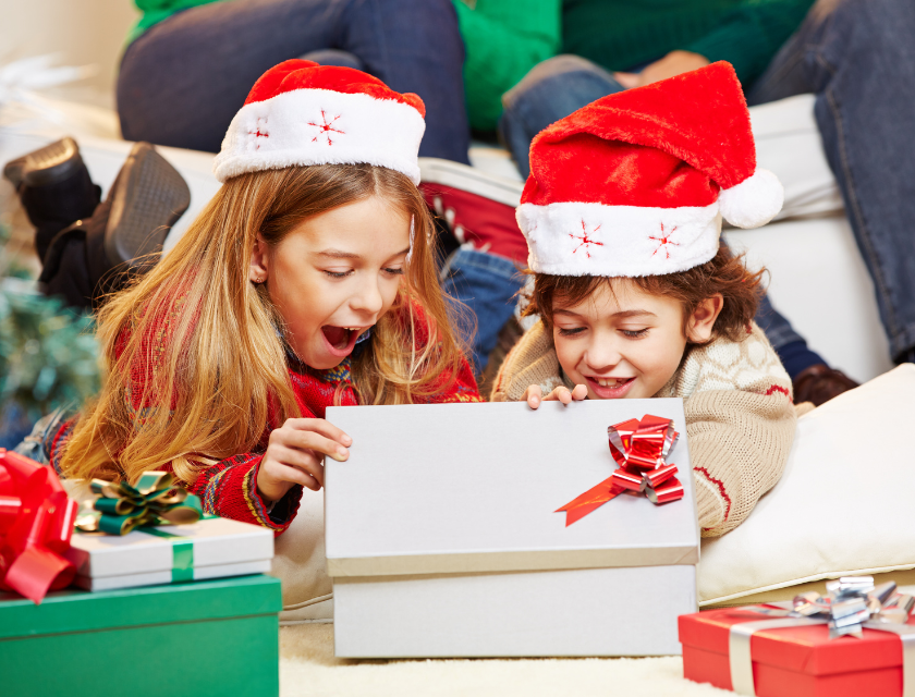 Two excited children wearing Santa hats opening a Christmas gift box. They are surrounded by festive presents.