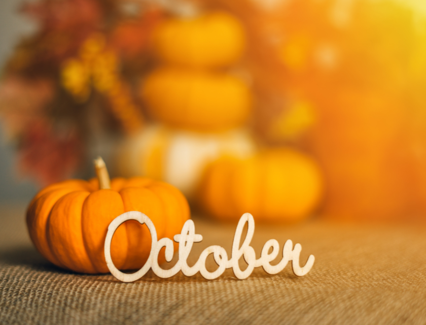 Small, bright orange pumpkin with a wooden "October" sign resting on a table, with a blurred background of autumn decorations.