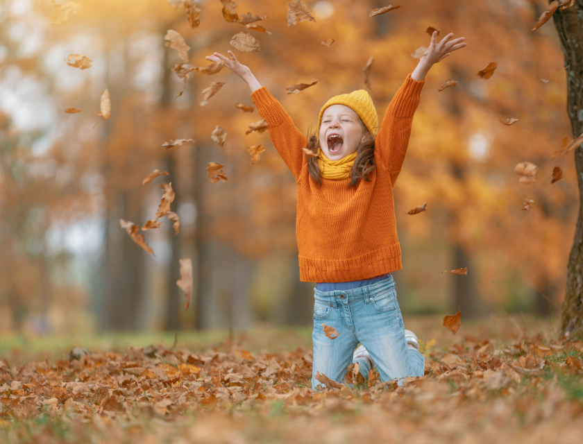 Child in an orange jumper and yellow hat throws dry autumn leaves in the air, surrounded by orange trees. Perfect for October half term fun in the countryside.