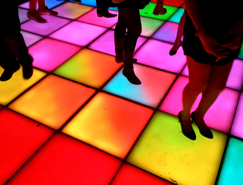 Bright, glowing squares of a dance floor light up in different colours as people dance at a disco party.