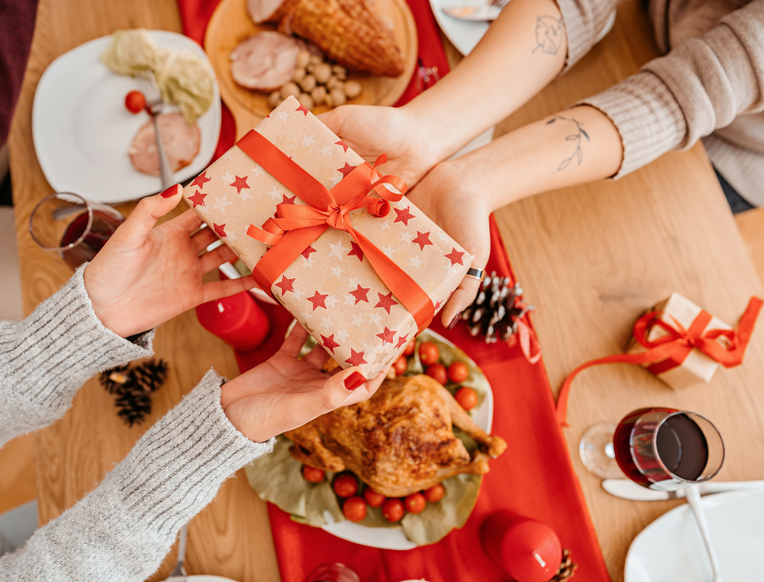 Two people exchanging a present wrapped in festive paper with red stars, sitting at a dinner table with turkey and decorations.