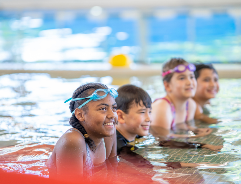 Four kids in swimsuits and goggles smile while having fun in an indoor swimming pool, ready to swim.