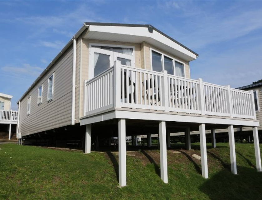 Beige static holiday caravan with large windows and a white-railed deck, lifted on stilts and set on a grassy slope under a clear blue sky. Ideal for your Haven Christmas break