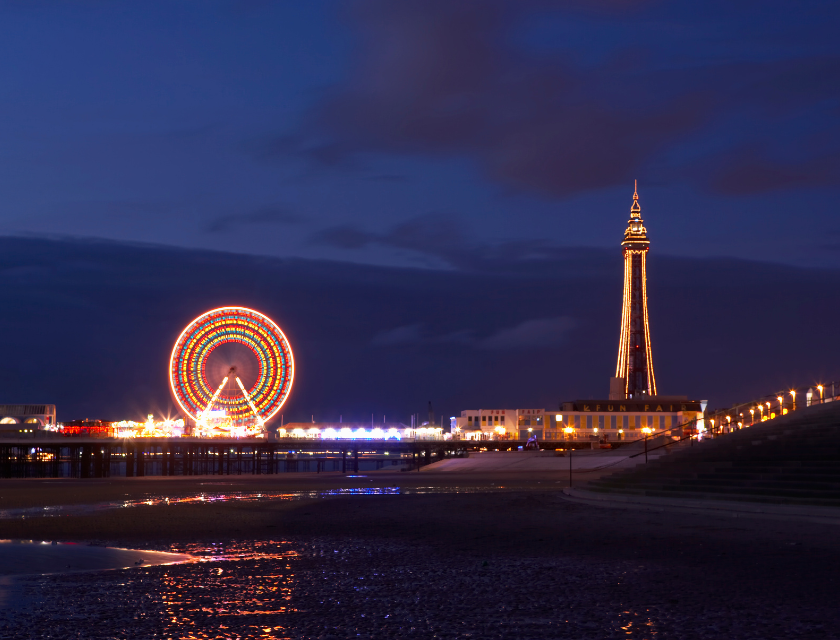 Blackpool Tower and a bright Ferris wheel are lit up in the evening, reflecting on the beach as the night sky darkens.