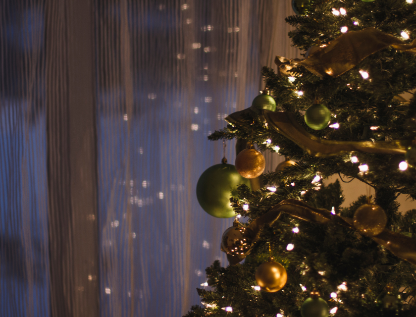 Decorated Christmas tree with green and gold baubles and glowing lights, standing next to a softly lit window.