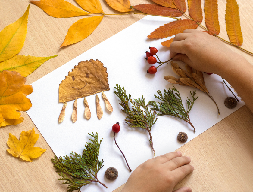 Child making an autumn craft by arranging leaves, twigs, and acorns on a piece of white paper. A perfect activity for autumn caravan breaks.