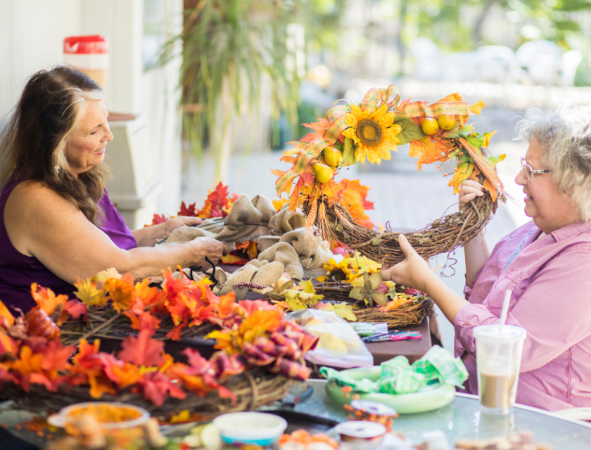 Two women sitting at a table, making an autumn wreath with sunflowers, leaves, and other autumn decorations.