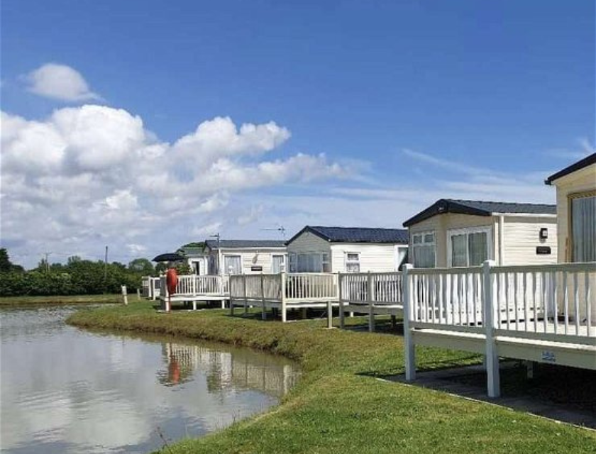 Static caravans with white fences, next to a calm lake under a blue sky, offering a peaceful UK autumn caravan activities in the countryside.