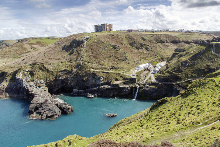 Tintagel, Cornwall, UK: April 14, 2016: A view of the main entrance to Tintagel Castle in the valley and Hotel Camelot on the cliff. There is a  beach cafe, visitors centre, public facilities and the booking desk and the entrance. The castle is maintained by English Heritage who are also responsible for Stonehenge and many other ancient sites across England.